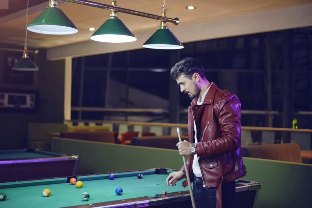 A man in a leather jacket holding a cue while lining up a shot on a billiards table.